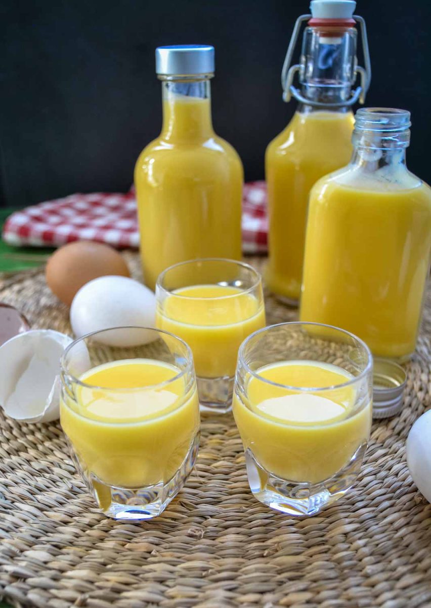 Cremiger Eierlikör selber machen im Glas mit Eiern und abgefüllten Flaschen im Hintergrund